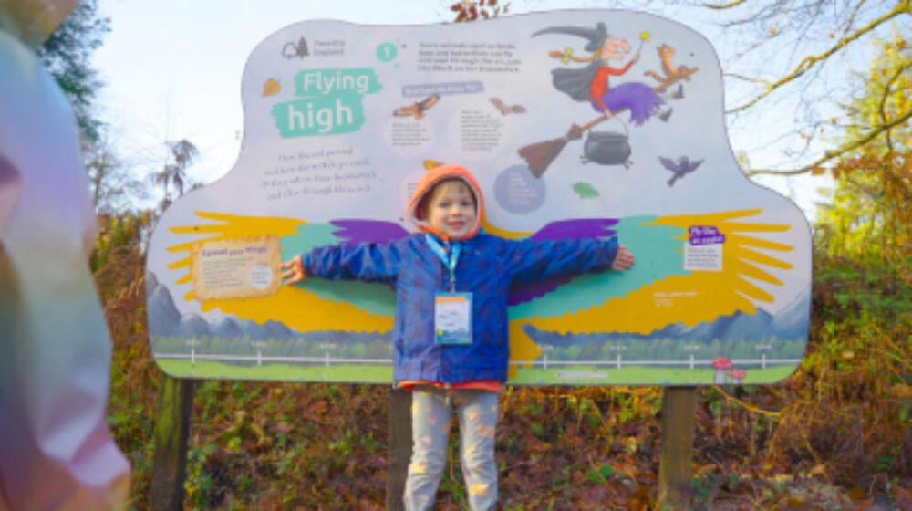 A child standing in front of a Room on the Broom trail panel at Fineshade Wood