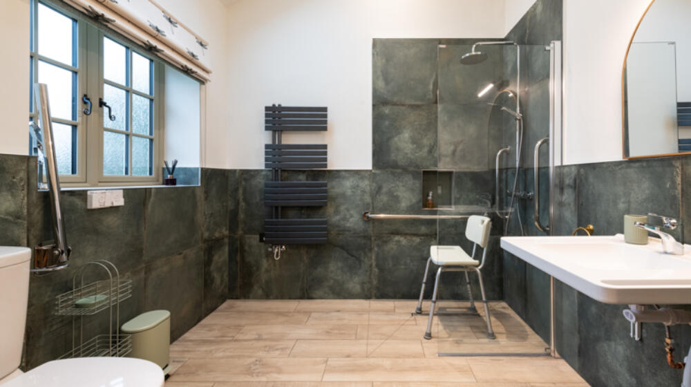 Accessible Wet room with non slip tiled floor, green tiled wall, moveable shower chair, glass shower screen, roll under sink, and comfort height WC